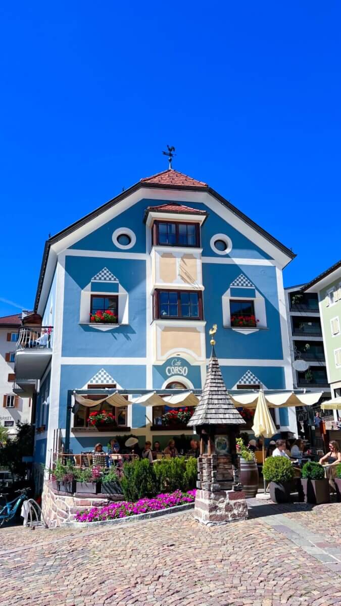 A Day in the Dolomites Italy: Ortisei, Karersee & Scenic Spots A bright blue Alpine-style building with white trim and red flower boxes, housing Café Corso, set on a cobblestone street under a vivid blue sky.