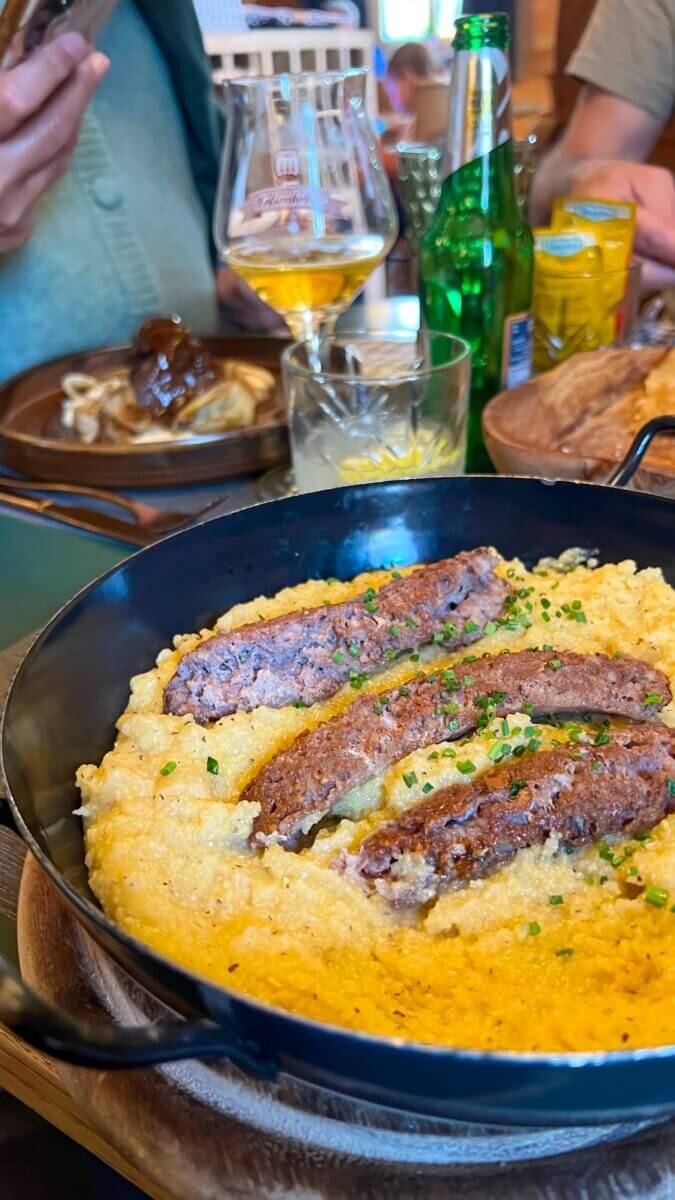 A Day in the Dolomites Italy: Ortisei, Karersee & Scenic Spots A close-up of a rustic meal showing grilled sausages served over creamy polenta in a black pan, with drinks and food on the table in the background.