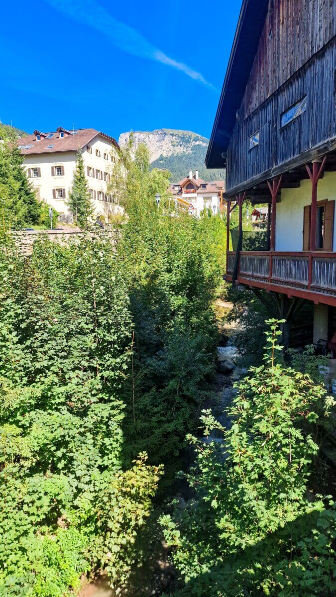 A Day in the Dolomites Italy: Ortisei, Karersee & Scenic Spots A lush green valley with dense trees, a small stream running through it, and Alpine-style houses in the background under a bright blue sky.