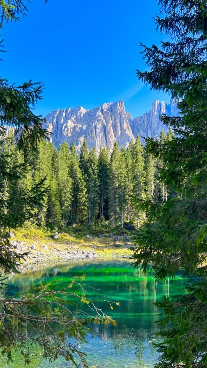 A Day in the Dolomites Italy: Ortisei, Karersee & Scenic Spots A clear green alpine lake surrounded by pine trees with the jagged peaks of the Dolomites rising in the background under a bright blue sky.