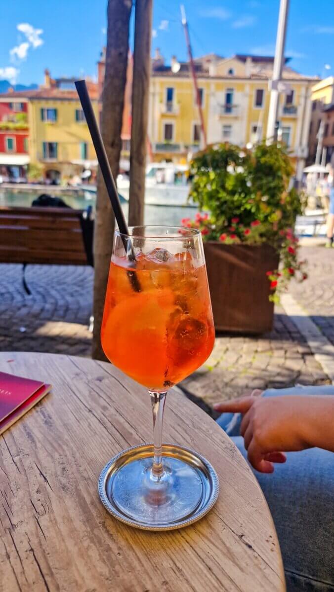 Discover the Most Beautiful Places Around Lake Garda Italy: Riva del Garda, Limone sul Garda, Malcesine and More Aperol Spritz on a café table in Sirmione, Italy, with pastel-colored buildings and the lake in the background.