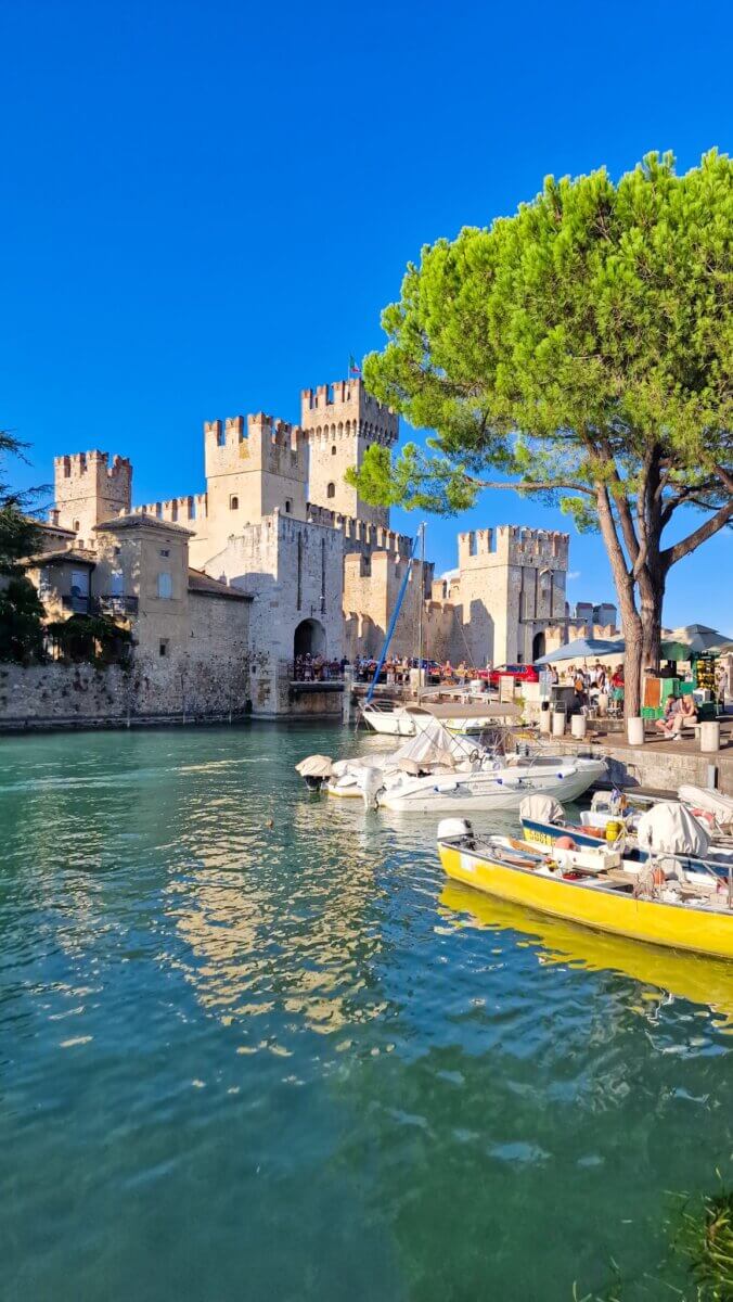 Discover the Most Beautiful Places Around Lake Garda Italy: Riva del Garda, Limone sul Garda, Malcesine and More Medieval castle with turquoise water and colourful boats in the foreground.
