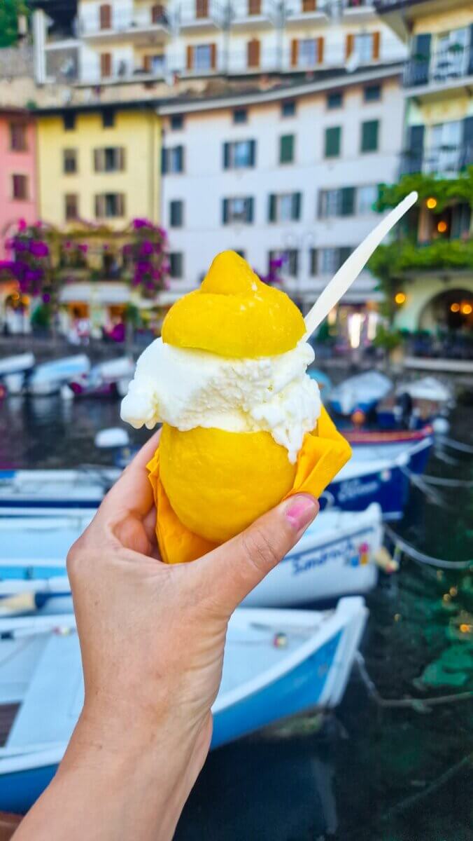 Discover the Most Beautiful Places Around Lake Garda Italy: Riva del Garda, Limone sul Garda, Malcesine and More Lemon sorbet served inside a frozen lemon shell in Limone sul Garda, Italy, with colorful lakeside buildings and boats in the background.
