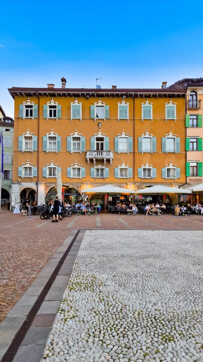 Discover the Most Beautiful Places Around Lake Garda Italy: Riva del Garda, Limone sul Garda, Malcesine and More Beautiful orange building with pastel blue shutters in a large square with cafés and restaurants