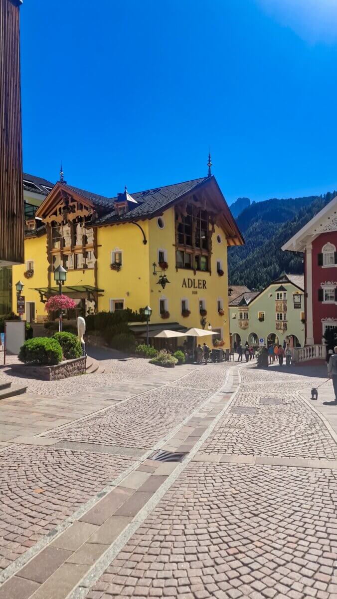 A Day in the Dolomites Italy: Ortisei, Karersee & Scenic Spots A sunny cobblestone square in a mountain village, featuring colorful Alpine buildings, including a yellow hotel labeled “Adler.”