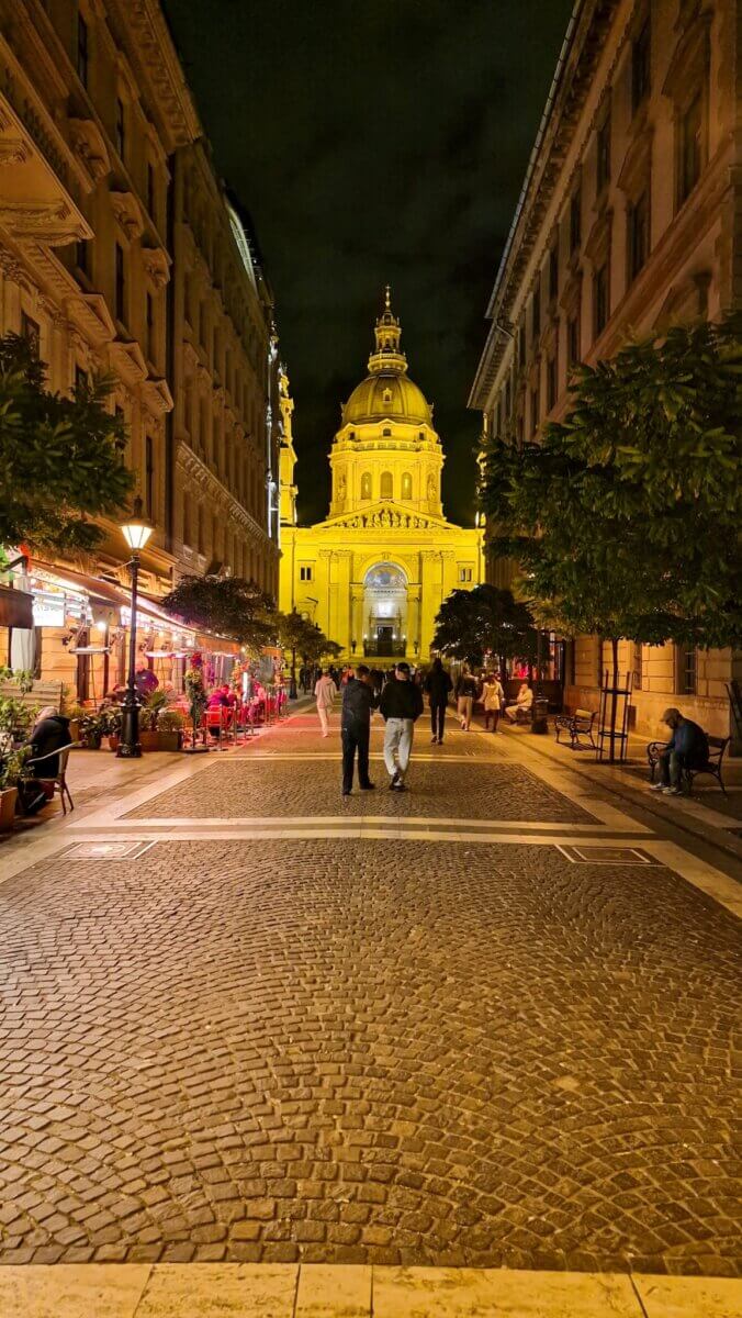 A cobblestone street at night, lined with lit cafes and trees, leads to a brightly illuminated historic building with a domed tower. People stroll leisurely, creating a warm, inviting ambiance.