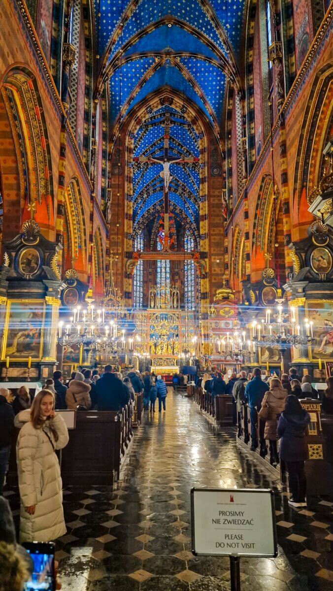 Interior of a large Gothic church filled with people during a service, with a bright blue vaulted ceiling decorated with gold stars, ornate arches, chandeliers, and a sign in the foreground that says “Please do not visit.”
