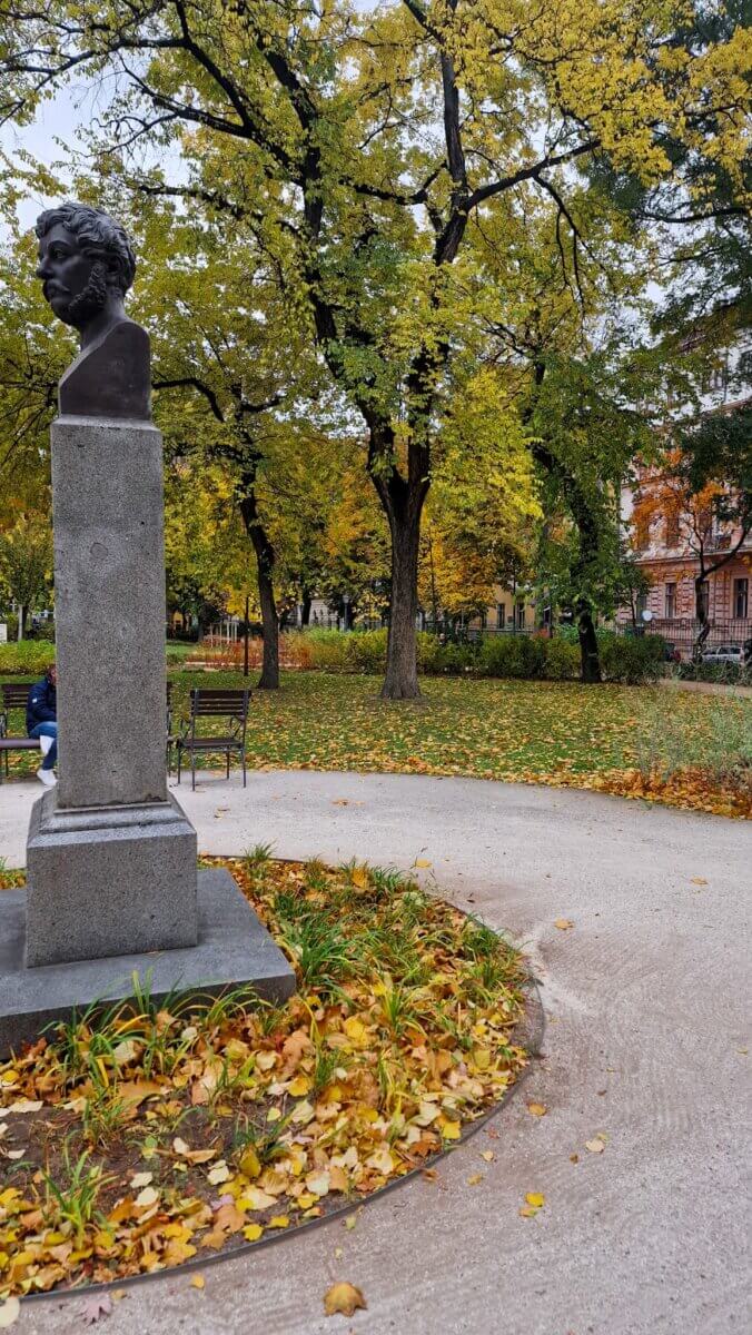 Bust statue on a pedestal in a park with autumn trees and fallen leaves. A person sits on a bench, creating a peaceful, reflective atmosphere.