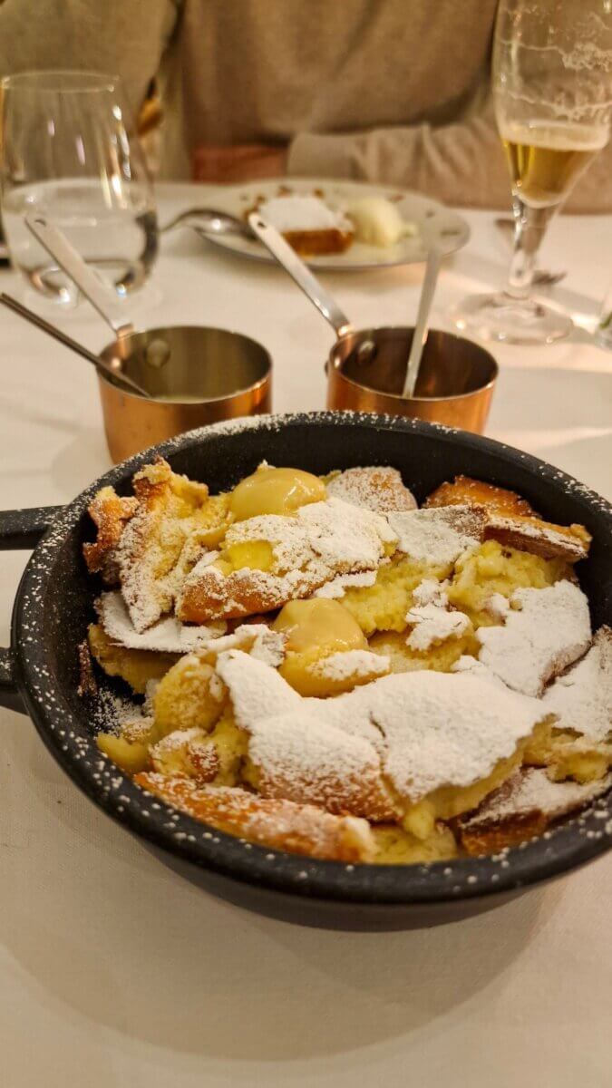 A cozy restaurant scene shows a cast-iron skillet with a dessert topped with powdered sugar and cream. Copper saucepans and drinks complete the table setting.