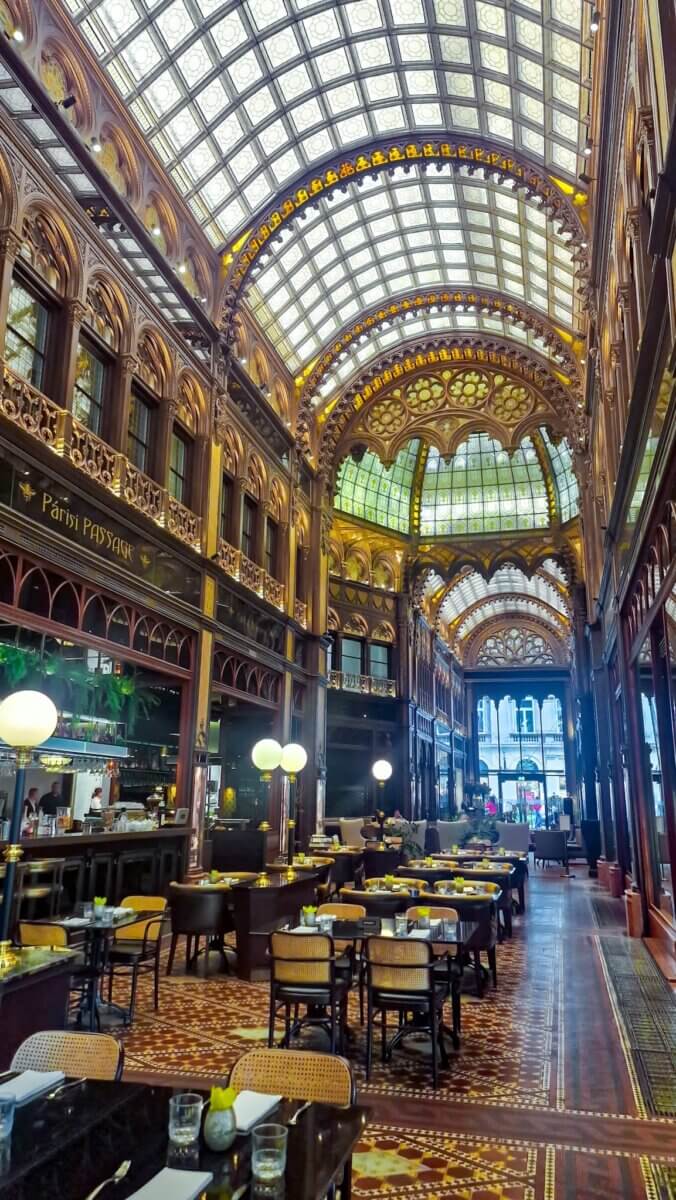 A grand restaurant with ornate architecture, high arched stained glass ceiling, patterned tile floors, and neatly set tables.