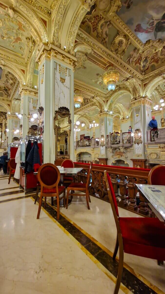 Luxurious cafe interior with ornate gold-accented arches, chandeliers, and red velvet chairs. The atmosphere is opulent and inviting.