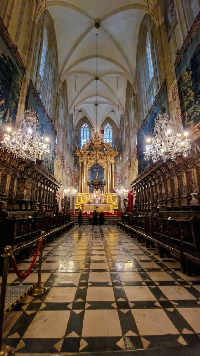 Interior of a tall, light-coloured church with vaulted ceilings, a golden altar at the front, chandeliers, and rows of carved wooden choir stalls lining both sides.