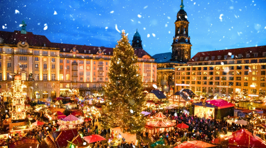 A bustling European Christmas market at night with a large decorated tree, festive stalls, lights, and snow falling over a historic town square.