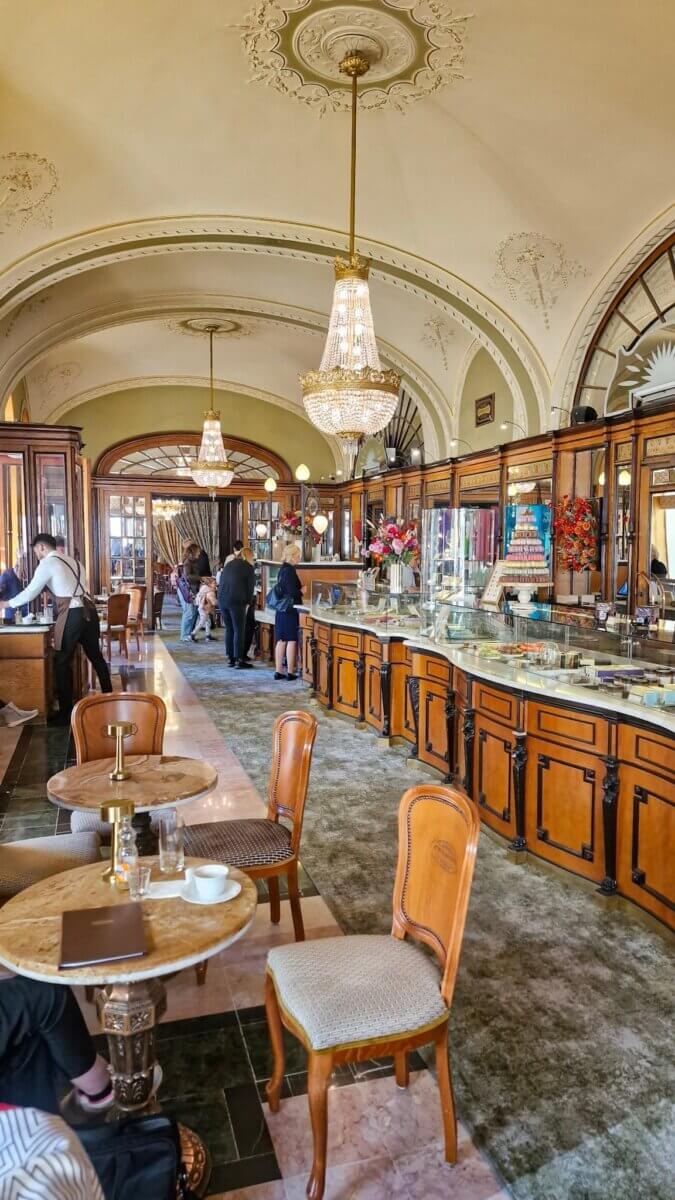 Elegant café interior with ornate chandeliers, arched ceilings, and wooden cabinets. Patrons converse at marble tables, creating a lively atmosphere.