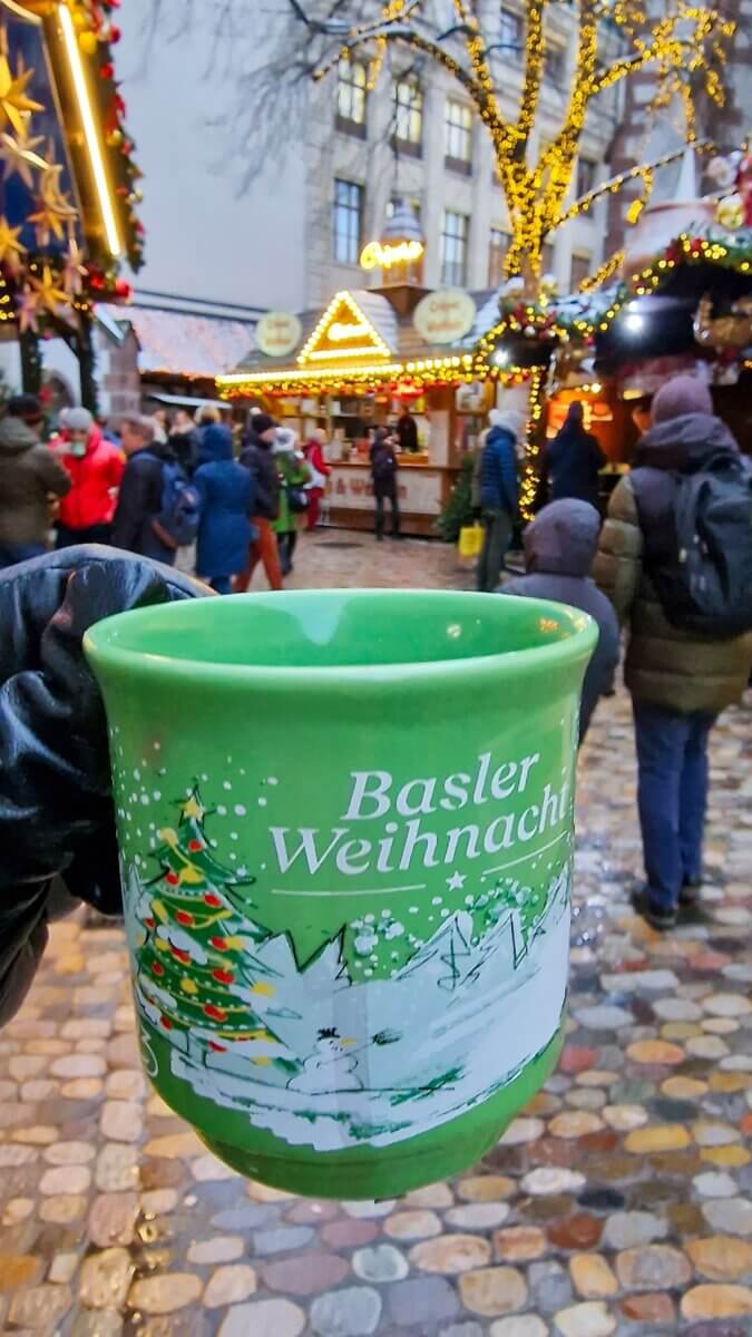 The image shows a green ceramic mug from the Basel Christmas market. The text "Basler Weihnacht" translates to "Basel Christmas" in English. The mug features a festive design with a Christmas tree and a snowman in a snowy landscape.