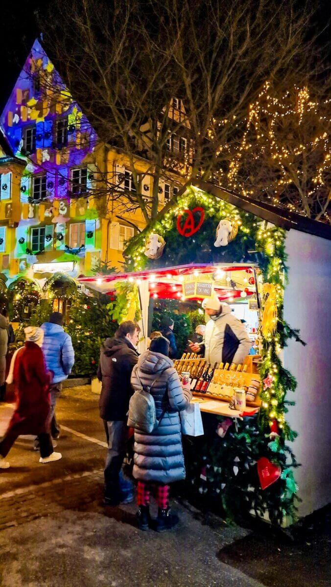 The image captures a lively scene at a Christmas market, likely in Europe, featuring a decorated stall and festive lighting.