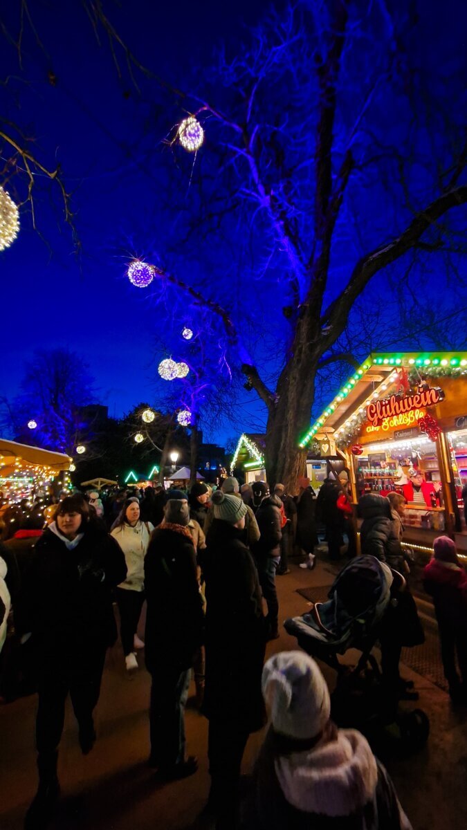 The image captures a lively scene at a Christmas market at night, featuring festive lighting and a stall selling Glühwein (mulled wine).