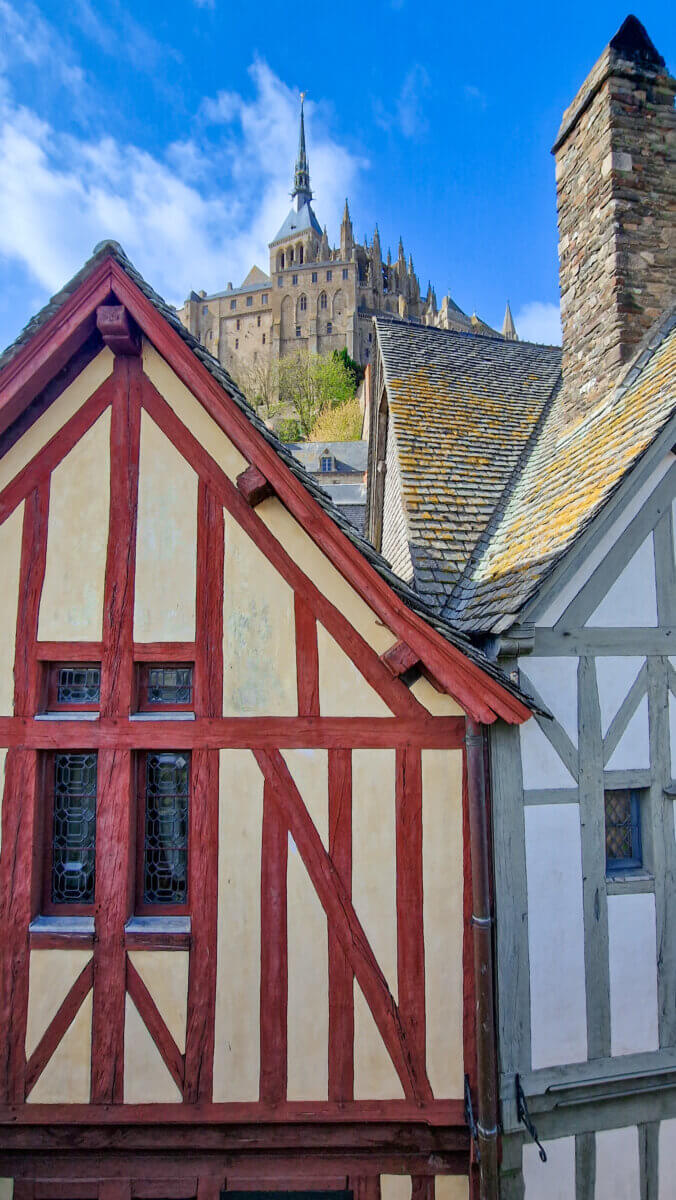 The image displays traditional half-timbered houses set against the Mont-Saint-Michel Abbey in Normandy, France
