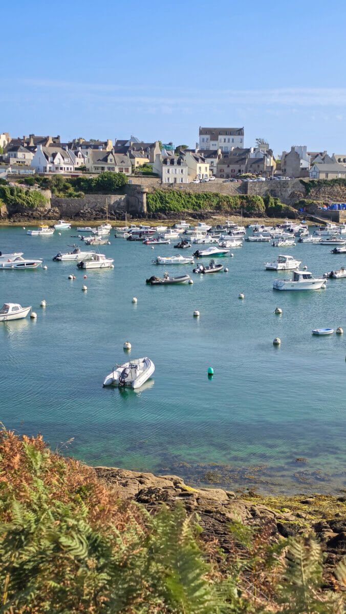 The image shows the scenic coastal harbor and town of Le Conquet in the Finistère department of Brittany, northwestern France, looking across the sheltered waters filled with moored boats toward the traditional stone houses and buildings lining the quay