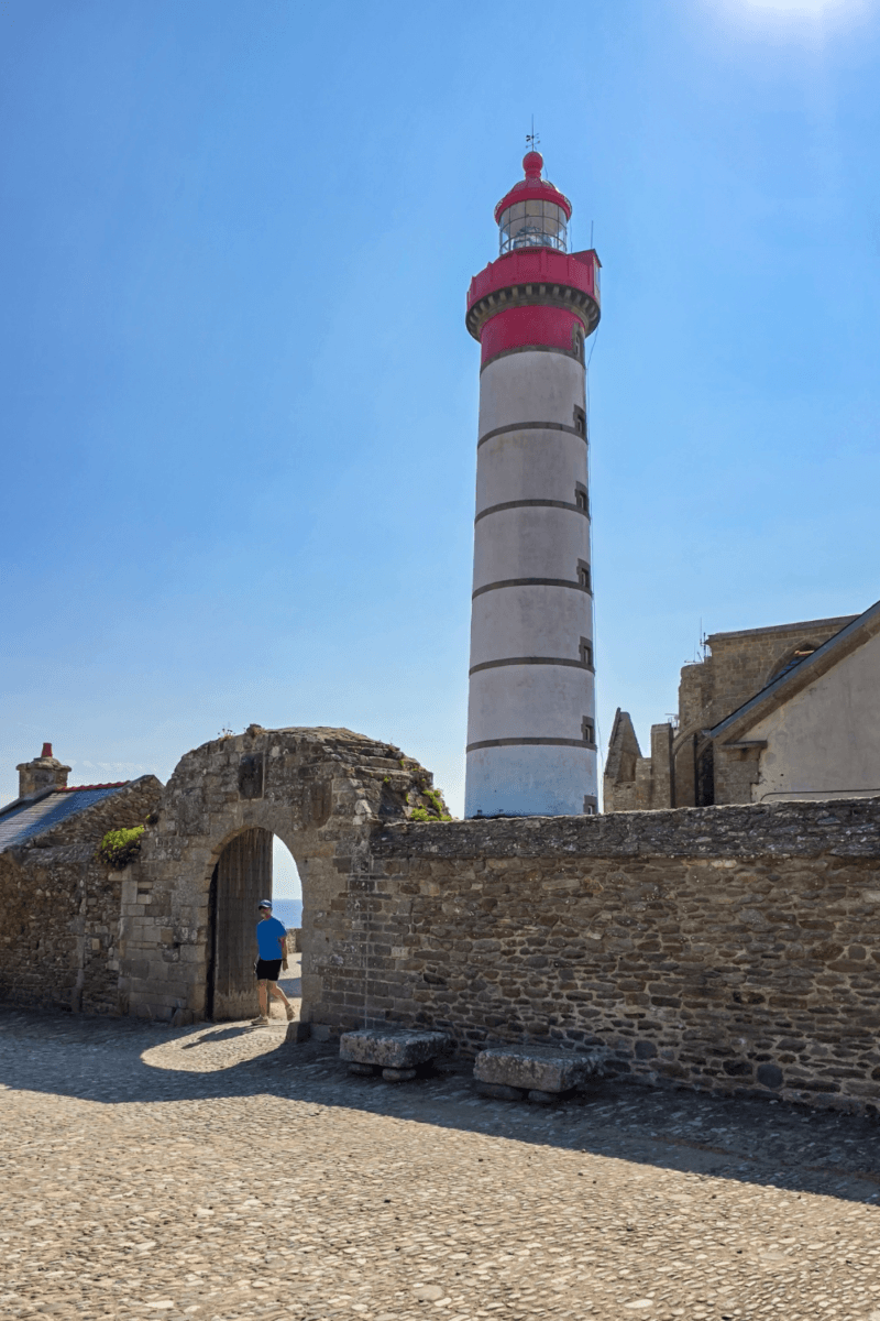 The image shows the historic Saint-Mathieu Lighthouse (Phare de Saint-Mathieu) alongside the stone walls and courtyard entrance near the medieval abbey ruins at Pointe Saint-Mathieu in Plougonvelin, Finistère, Brittany, France