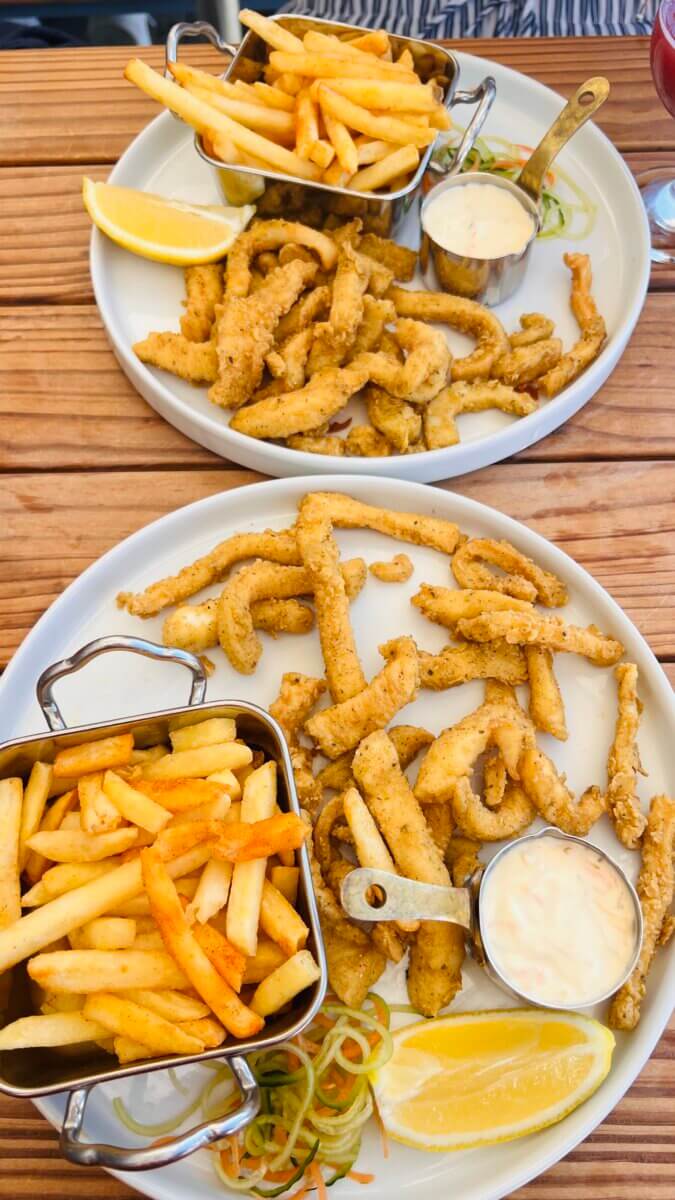 Crispy fried calamari or squid, golden french fries in a metal basket, a small pot of creamy dipping sauce, and a side of shredded salad.