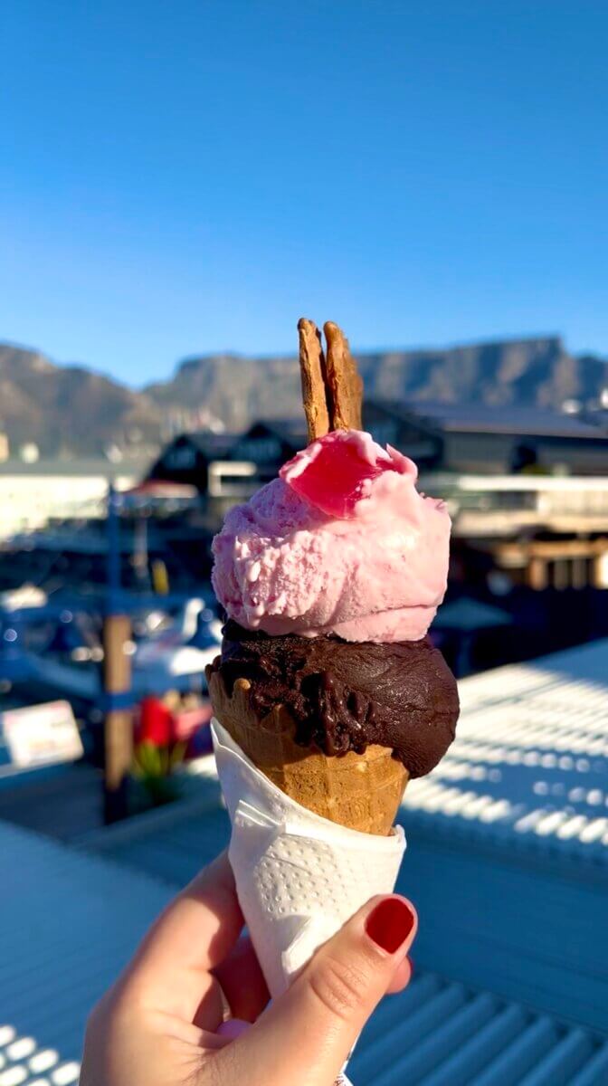 The image shows an ice cream cone with a scoop of dark chocolate gelato on the bottom, a scoop of pink Turkish delight gelato on top, and a biscuit or wafer stuck in it.