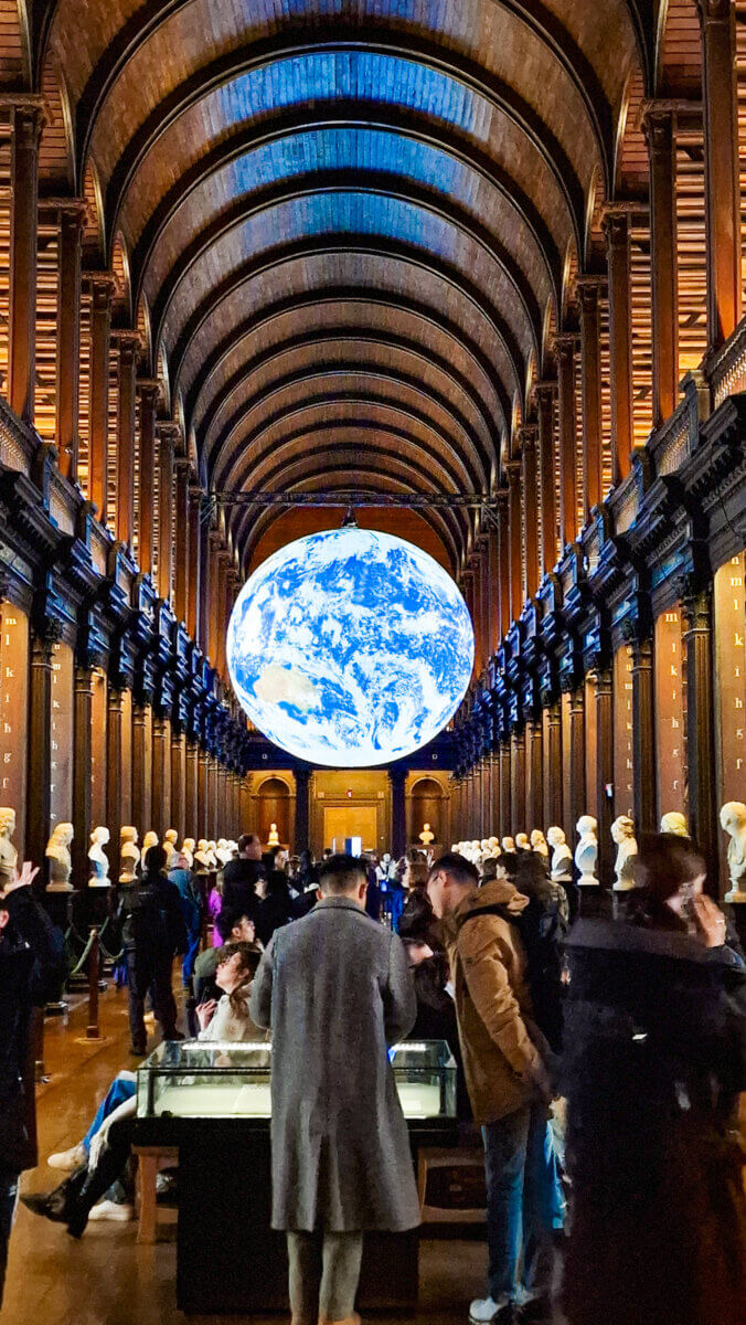 The image shows the famous Long Room of the Old Library at Trinity College Dublin in Dublin, Ireland, featuring the illuminated floating globe installation titled Gaia by artist Luke Jerram.