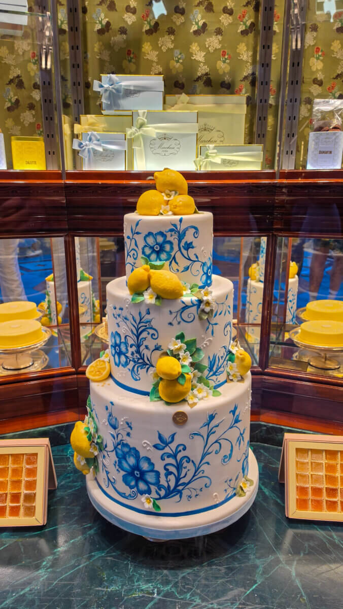The image shows a three-tiered decorative cake with a white fondant finish, hand-painted blue floral motifs, and modeled yellow lemons